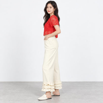 A woman models a red lace top, cream ruffled pants, and white loafers against a studio backdrop.