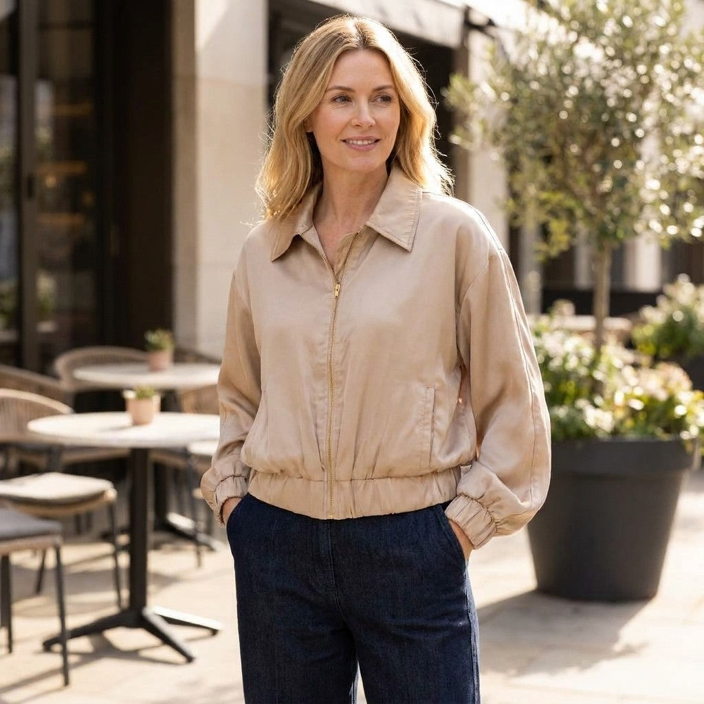 A woman in a beige jacket and dark trousers stands outside a sunlit café.