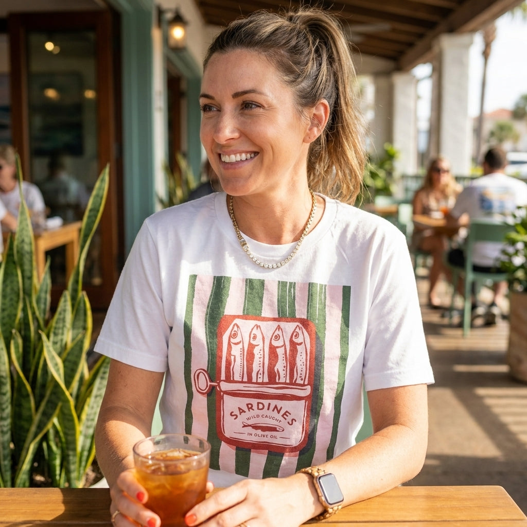 Smiling woman with iced tea, wearing a sardine-themed shirt at a sunny outdoor café.