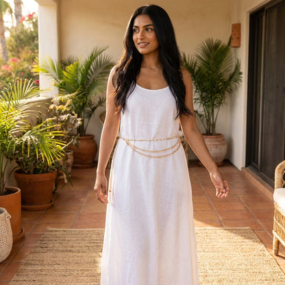 Woman in a white dress with gold chain, in a sunny, plant-filled room.