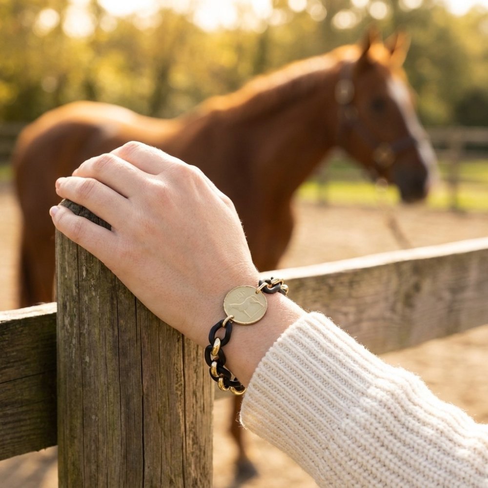 A hand wearing a horse-themed bracelet rests on a wooden fence, with a horse in the background.