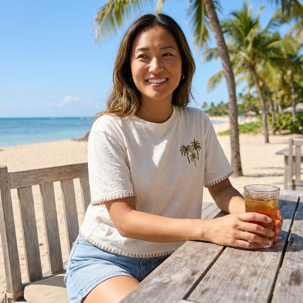 Smiling woman with iced tea at a beachside table under palm trees.