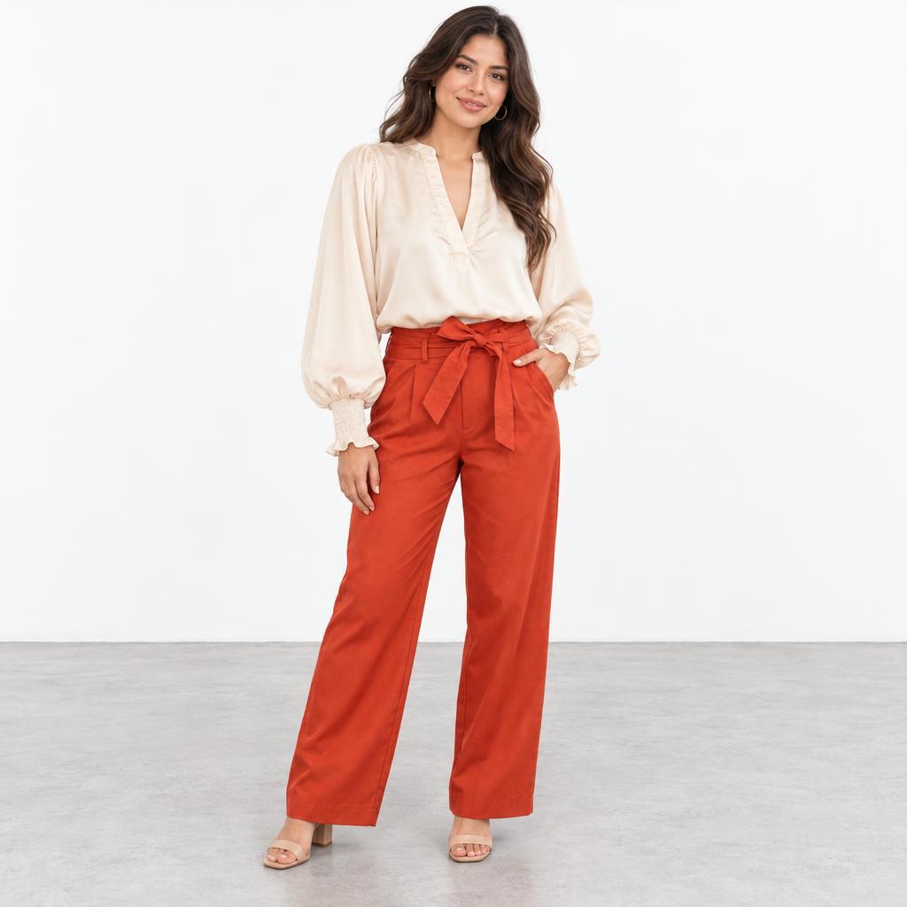 A woman models a cream blouse and rust-red, high-waisted trousers against a plain backdrop.