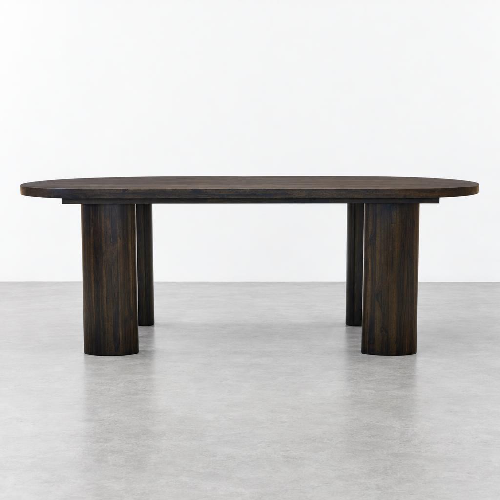 A dark wooden oval dining table with sturdy cylindrical legs in a minimalist studio.