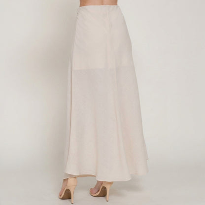 Woman in a long white skirt and nude heels, standing on a plain background.