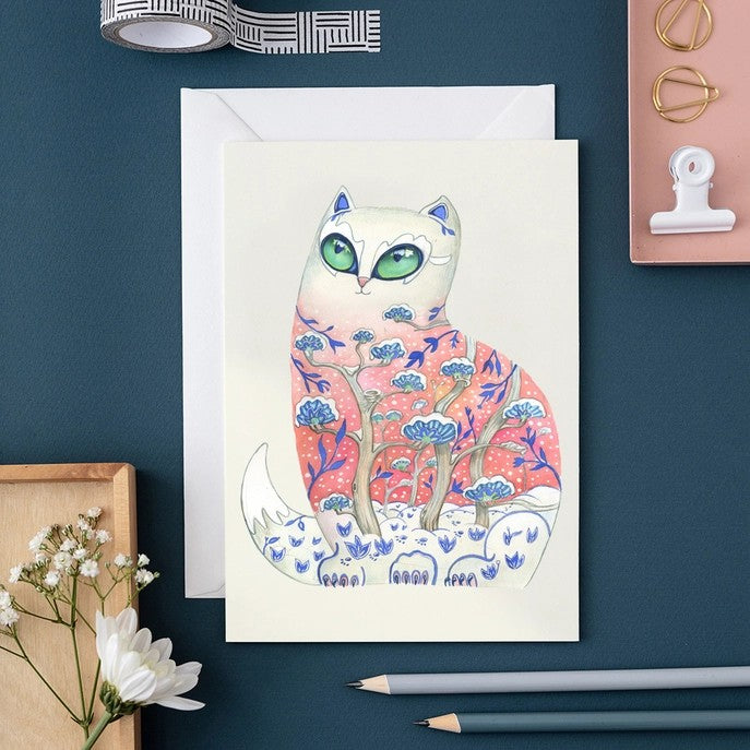 Whimsical cat art card on a desk with pencils and flowers.