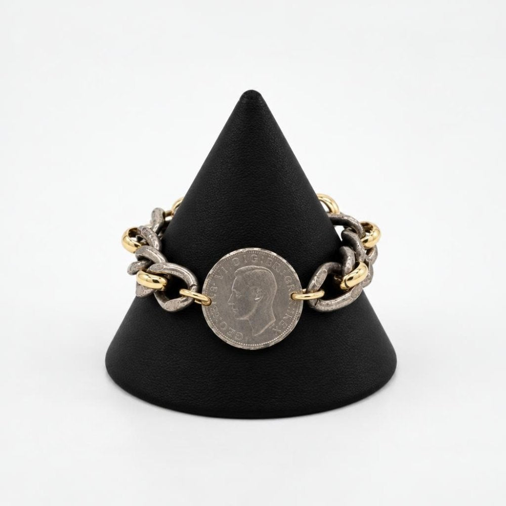A mixed-metal chain bracelet with a vintage coin displayed on a black stand.