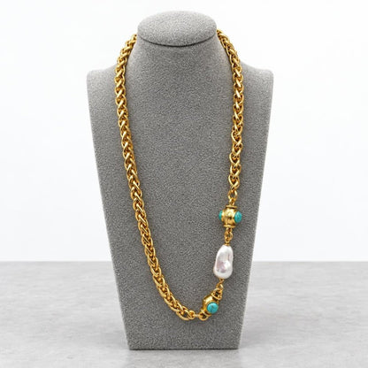 Gold chain necklace with turquoise accents and a pearl, displayed on a gray stand.