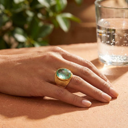 A hand wearing a gold ring with a large green gemstone rests on a sunlit table.