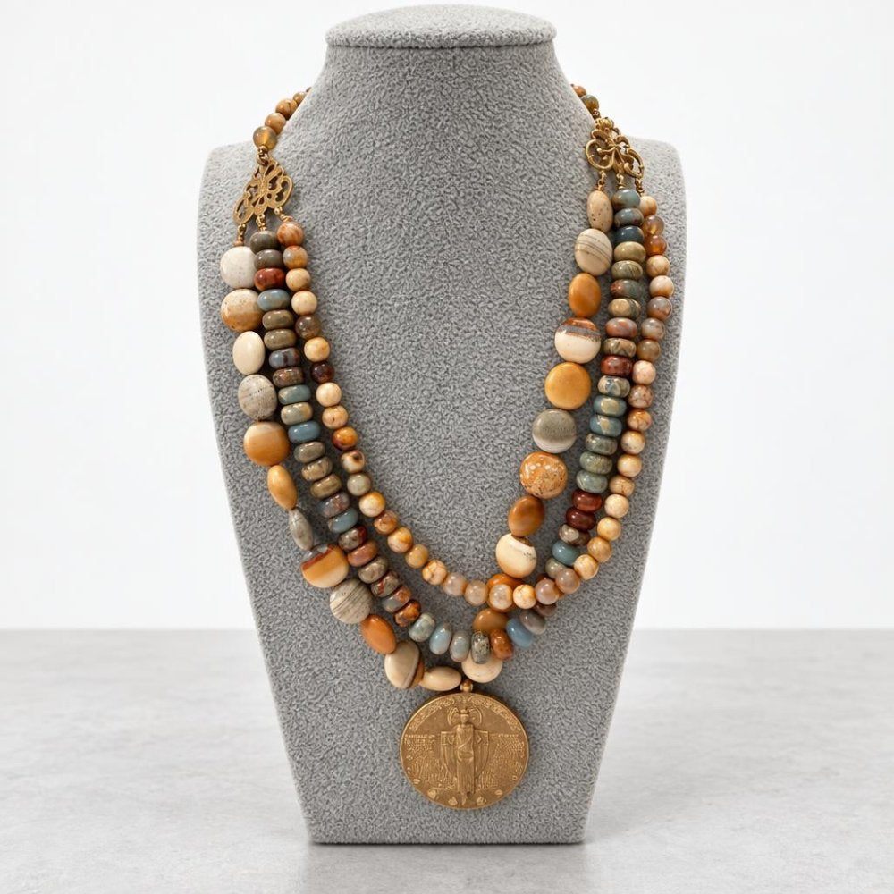 A layered necklace of earthy stone beads with a large bronze medallion, displayed on a gray bust.