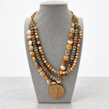A layered necklace of earthy stone beads with a large bronze medallion, displayed on a gray bust.