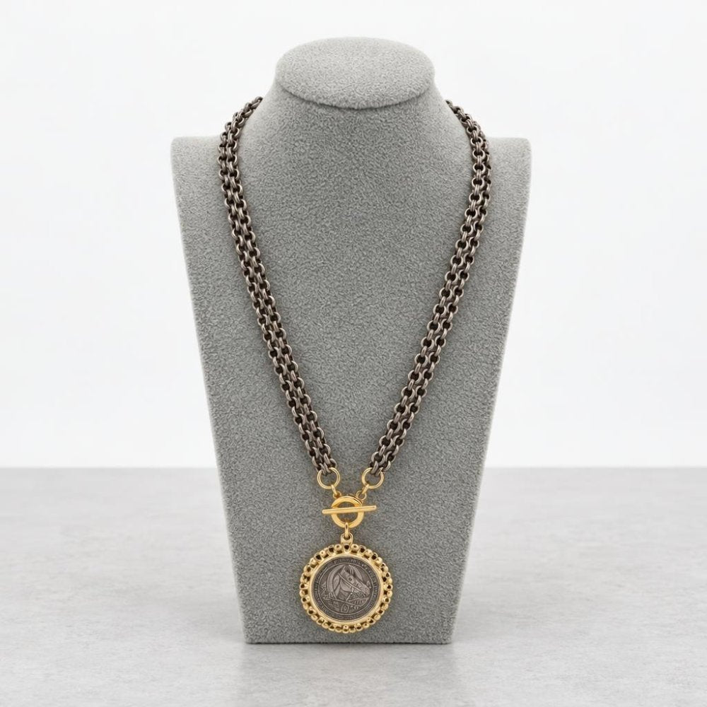 A dark metal chain necklace with a gold-toned medallion pendant on a gray display bust.