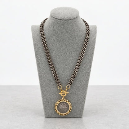 A dark metal chain necklace with a gold-toned medallion pendant on a gray display bust.