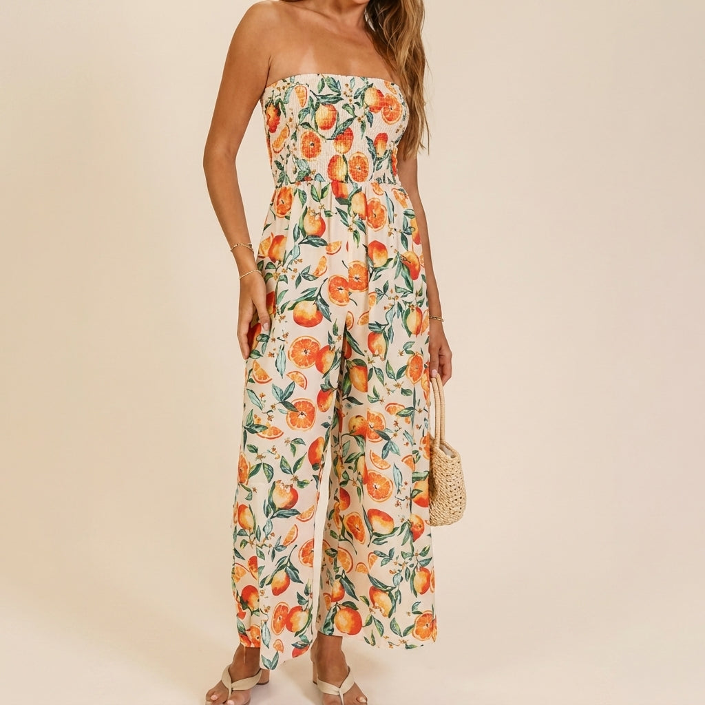 Model in a strapless, orange fruit-patterned jumpsuit holding a woven bag.