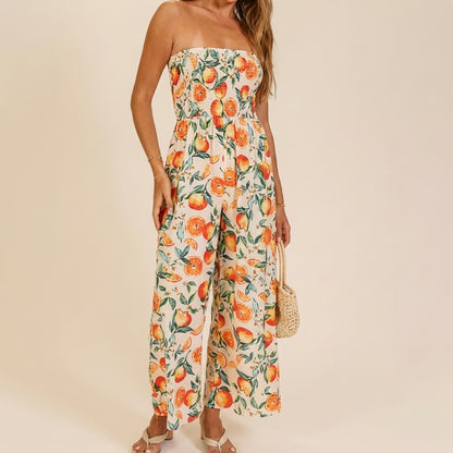 Model in a strapless, orange fruit-patterned jumpsuit holding a woven bag.