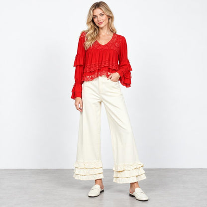Model wears a red lace-trimmed blouse with ruffled sleeves, cream wide-leg pants, and white loafers.