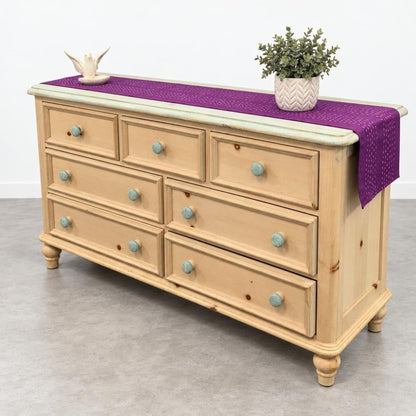 A light wood dresser with turquoise knobs, topped with a purple runner and potted plant.