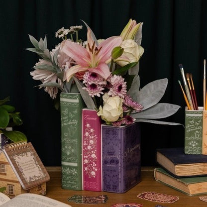 Vases shaped like books hold flowers beside stacked books and art supplies on a desk.
