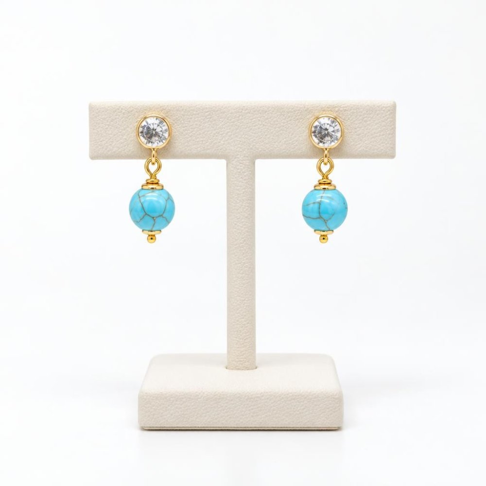 Gold drop earrings with clear gemstones and turquoise beads displayed on a stand.