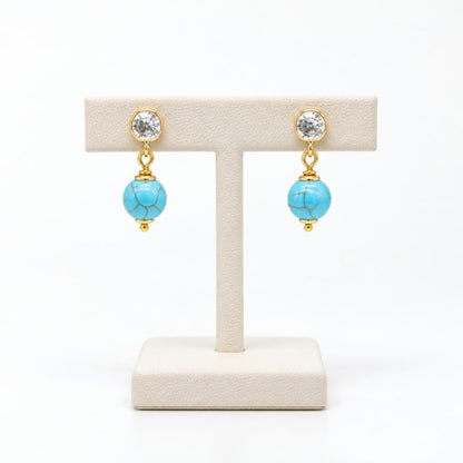 Gold drop earrings with clear gemstones and turquoise beads displayed on a stand.