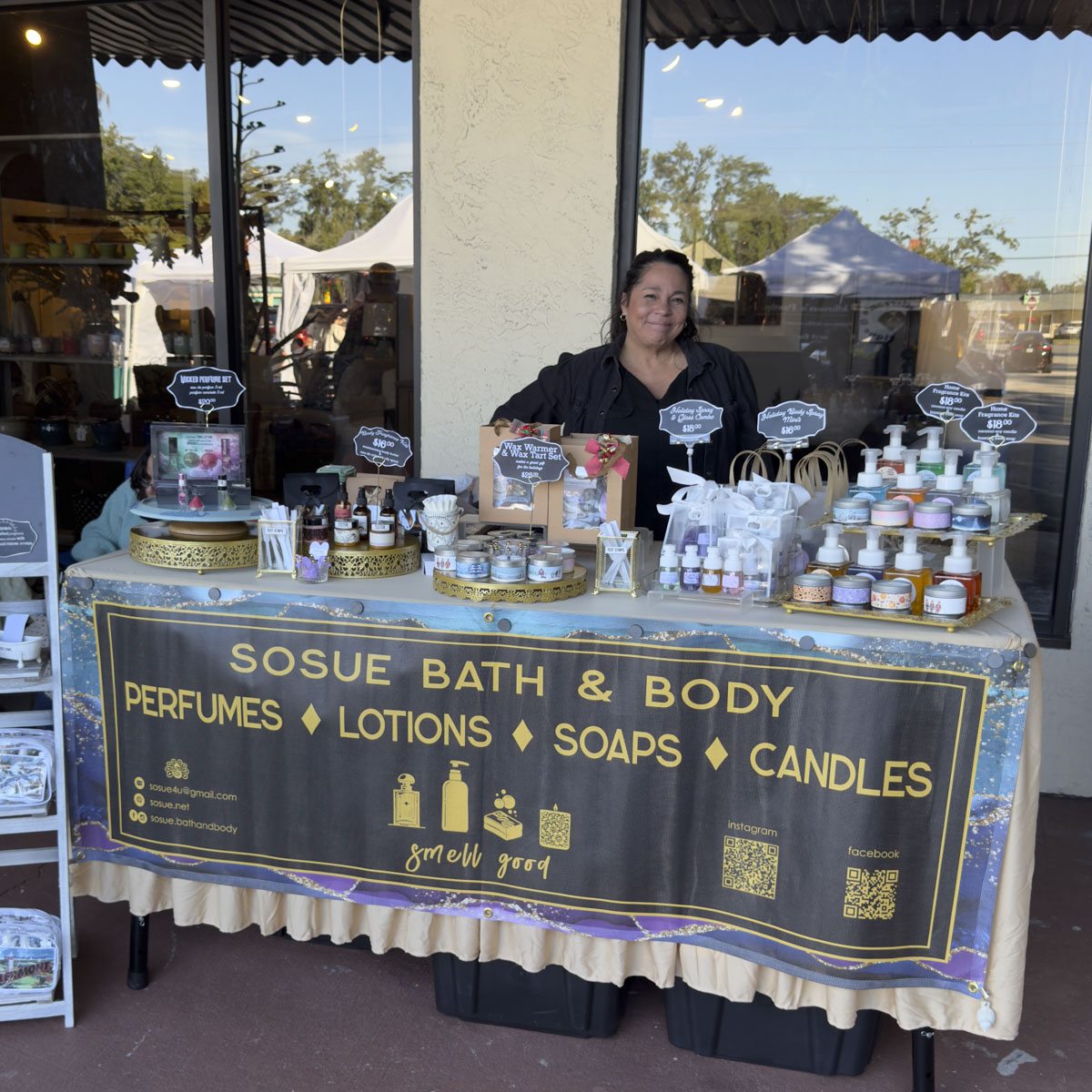A bath and body vendor at a table under the overhang, with market tents behind